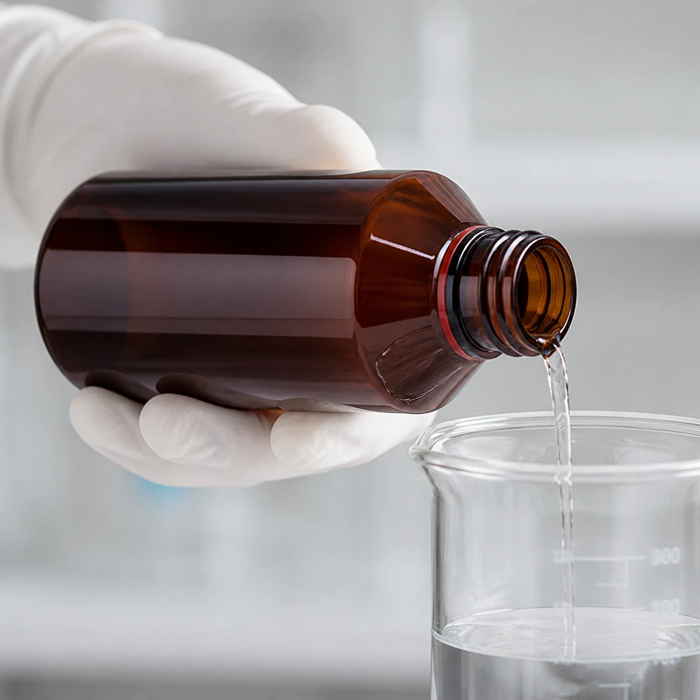 Ruka v rukavici nalévá čirou tekutinu z jantarové PET láhve o objemu 200 ml do skleněné kádinky v laboratoři
