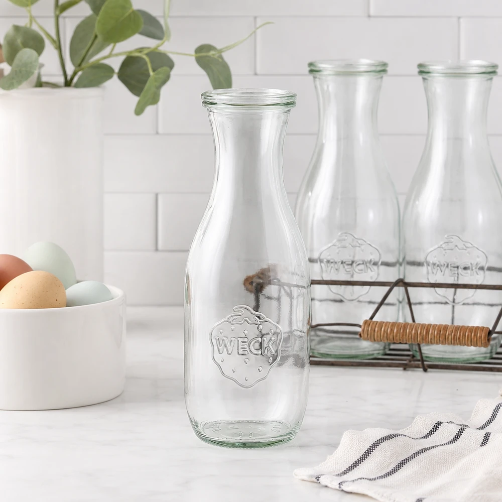 1062ml láhev na šťávu se skleněným víčkem WECK RR60 WECK Saftflasche 1062ml Glasflasche leer in moderner weißer Küche mit Eierschale Drahtkorb und Pflanze