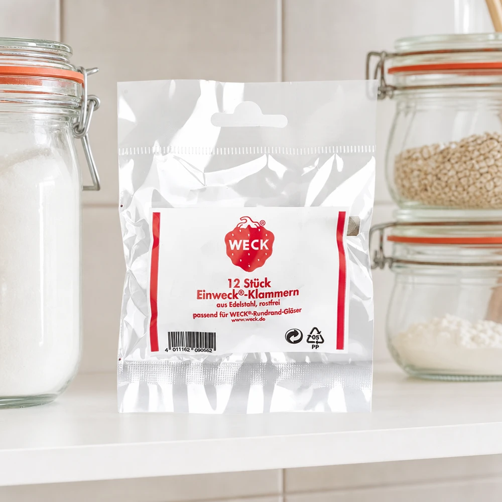 Svorky Original WECK (balení s 12 kusy) WECK Klammern Produktverpackung in Küche zwischen Vorratsgläsern mit Getreide Mehl auf weißer Arbeitsplatte Küchenambien