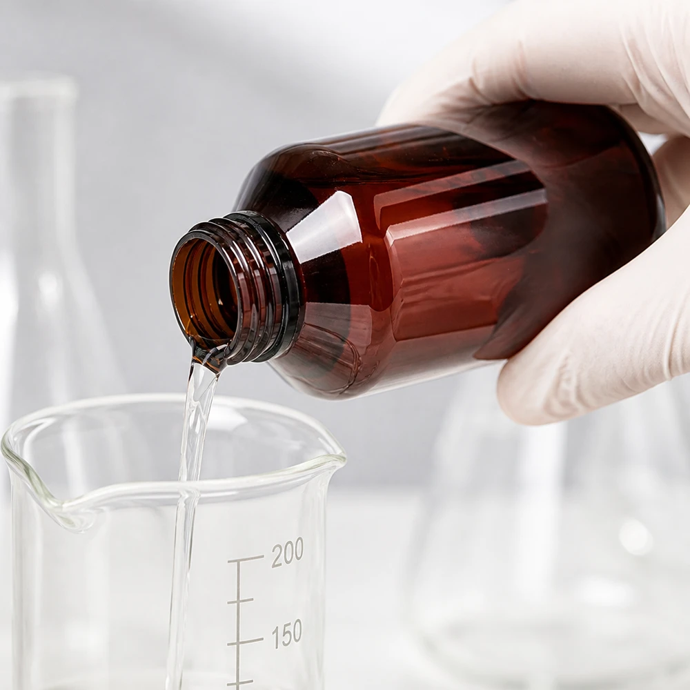 PET láhev 100 ml s bílým/červeným uzávěrem s ochranou proti manipulaci Čirá tekutina se nalévá z hnědé PET láhve o objemu 100 ml do skleněné kádinky